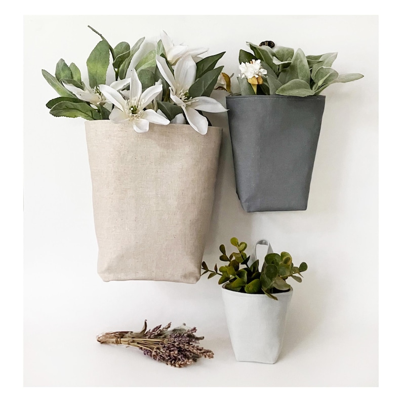 May include: Three fabric wall planters in beige, gray, and white, each filled with greenery and flowers. A bundle of dried lavender sits below. The planters are designed to hang on a wall, adding a touch of nature to any space.