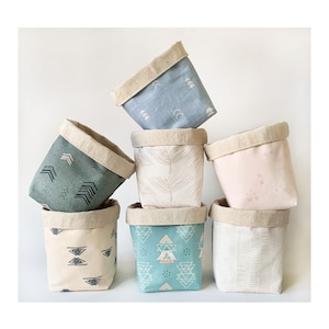 May include: Seven fabric storage baskets in a stack, in shades of blue, green, pink, and cream. Each basket has a different geometric or botanical pattern and a natural linen rim. These baskets are designed for storage and organization.