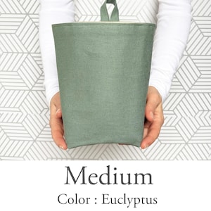 May include: A medium-sized, eucalyptus-colored fabric container with a loop handle. The container is cylindrical with a folded top edge and a light-colored interior. The text "Medium" and "Color: Eucalyptus" are displayed below the container.