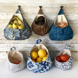 Puede incluir: Seis cestas de almacenamiento de tela colgantes con diferentes patrones y colores. Las cestas est&aacute;n llenas de varias frutas y verduras, incluyendo pl&aacute;tanos, limones, limas, aguacates y tomates.