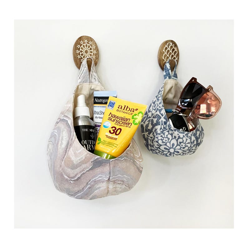 Puede incluir: Dos bolsillos de pared de tela con ganchos de madera, uno con un patr&oacute;n de m&aacute;rmol marr&oacute;n claro y el otro con un patr&oacute;n floral azul y blanco. El bolsillo con el patr&oacute;n de m&aacute;rmol contiene una botella de protector solar, una botella de spray y una peque&ntilde;a botella de loci&oacute;n. El bolsillo con el patr&oacute;n floral contiene un par de gafas de sol.