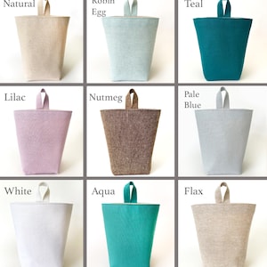 May include: Nine fabric doorstops in various colors, including natural, robin egg, teal, lilac, nutmeg, pale blue, white, aqua, and flax. Each doorstop has a trapezoidal shape with a small loop handle at the top.