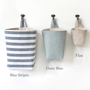 May include: Three hanging fabric storage baskets in blue stripes, dusty blue, and flax colors. Each basket has a loop for hanging and a neutral-colored interior. The baskets are labeled "Blue Stripes", "Dusty Blue", and "Flax".