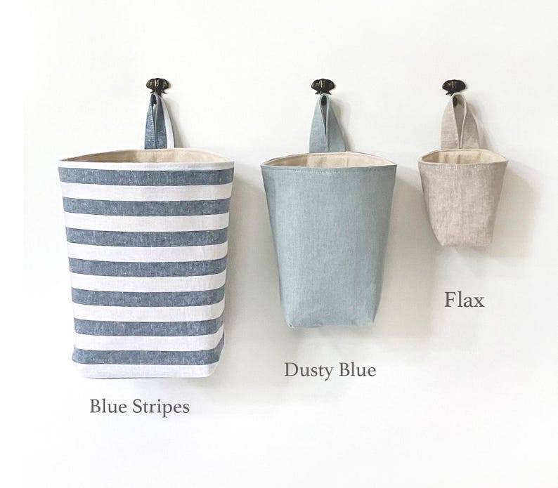May include: Three hanging fabric storage baskets in blue stripes, dusty blue, and flax colors. Each basket has a loop for hanging and a neutral-colored interior. The baskets are labeled "Blue Stripes", "Dusty Blue", and "Flax".