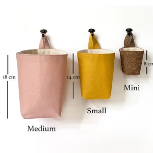 May include: Three hanging fabric storage baskets in varying sizes and colors. The largest is a medium pink, 18 cm tall. The small is mustard yellow, 14 cm tall. The mini is brown, 8 cm tall. Each has a loop for hanging.