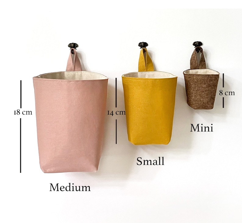 May include: Three hanging fabric storage baskets in varying sizes and colors. The largest is a medium pink, 18 cm tall. The small is mustard yellow, 14 cm tall. The mini is brown, 8 cm tall. Each has a loop for hanging.