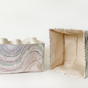 May include: Two fabric storage bins. One bin is filled with white spools of thread. The other bin is empty. The bins have a square shape. One bin has a marbled pattern in shades of purple, green, and tan. The other bin has a gray and white floral pattern.
