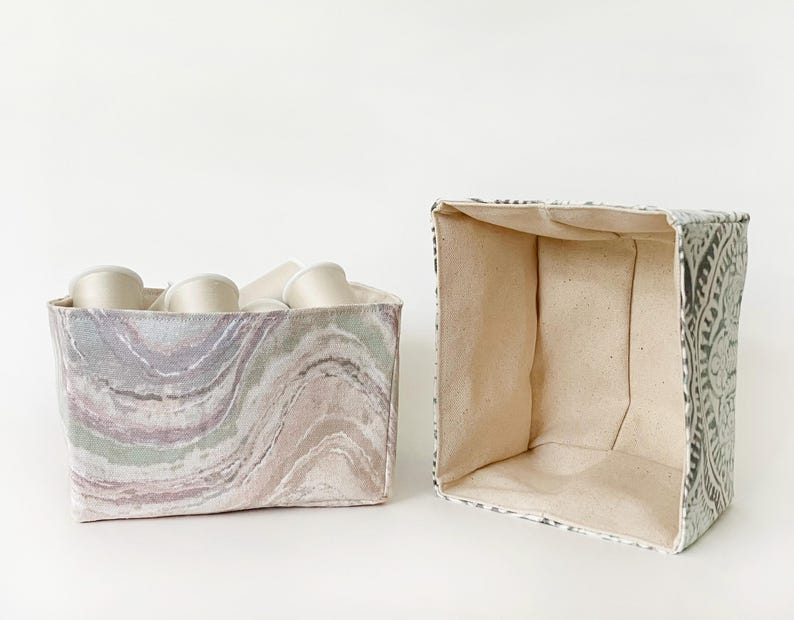 May include: Two fabric storage bins. One bin is filled with white spools of thread. The other bin is empty. The bins have a square shape. One bin has a marbled pattern in shades of purple, green, and tan. The other bin has a gray and white floral pattern.