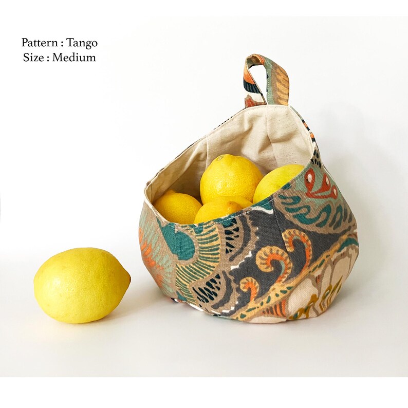 Puede incluir: Una cesta de tela colgante con un estampado paisley colorido en tonos marrones, verdes y naranjas. La cesta est&aacute; llena de limones y tiene un forro de color crema. El texto "Pattern: Tango Size: Medium" es visible en la esquina superior izquierda de la imagen.