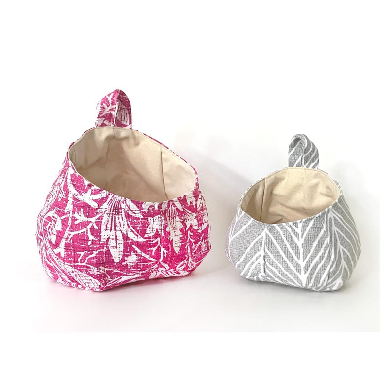 Puede incluir: Dos cestas colgantes de tela, una rosa con un patr&oacute;n floral blanco y otra gris con un patr&oacute;n de chevron blanco.