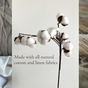 May include: A sprig of cotton with fluffy white blooms and brown stems, set against a neutral background. The image includes the text "Made with all-natural cotton and linen fabrics."
