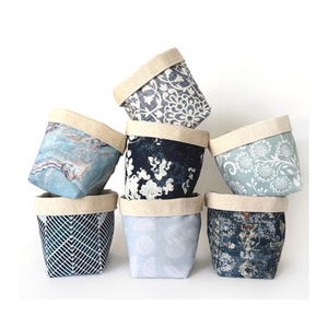 Puede incluir: Una colección de siete cestas de almacenamiento de tela con varios patrones y colores. Las cestas presentan un borde de color neutro y una variedad de diseños, incluyendo patrones florales, geométricos y abstractos en tonos de azul, blanco y gris.