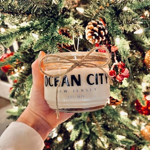 May include: A white candle jar with a brown twine bow and a black label that reads "Ocean City, New Jersey, Est. 1879". The candle jar is in front of a Christmas tree with red and silver ornaments.