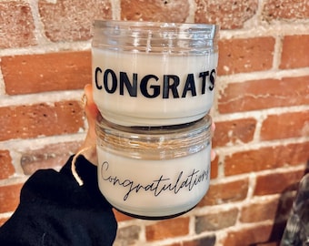 May include: Two glass jar candles with white wax. The top candle has the word "CONGRATS" in black block letters. The bottom candle has the word "Congratulations" in a cursive font. The background is a red brick wall.