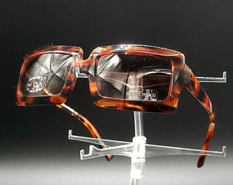 May include: Brown and tortoise shell square-shaped sunglasses with a clear lens. The sunglasses are on a clear plastic display stand.