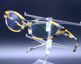 May include: A pair of black and yellow tortoise shell eyeglasses with green lenses. The glasses are on a clear plastic display stand.