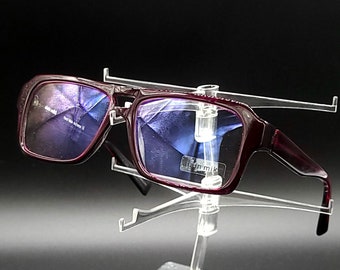 May include: Burgundy-colored rectangular eyeglasses with blue tinted lenses. The glasses are on a clear plastic display stand.