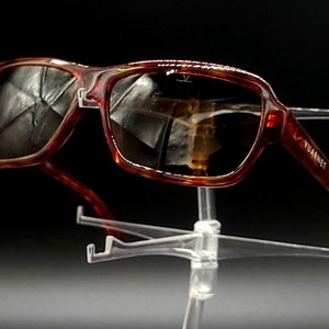 May include: A pair of brown tortoise shell sunglasses with a dark lens. The sunglasses are on a clear plastic display stand. The brand name "Vuarnet" is printed on the right arm of the sunglasses.