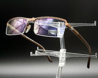 May include: Brown metal frame eyeglasses with clear lenses. The glasses are on a clear plastic display stand. The brand name "SHARIK" is visible on the right lens.