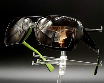 May include: Black sunglasses with green arms and brown lenses. The sunglasses are on a clear plastic stand.