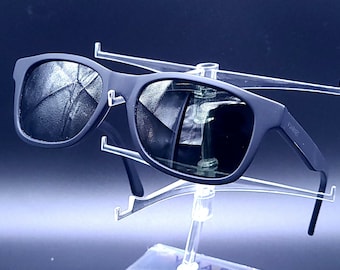 May include: Black sunglasses with a square frame and dark lenses. The sunglasses are displayed on a clear plastic stand.