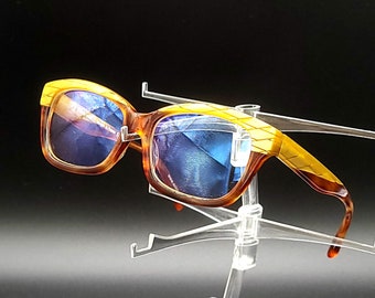May include: A pair of brown and yellow tortoise shell sunglasses with blue lenses. The sunglasses are on a clear plastic display stand.