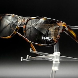 Puede incluir: Gafas de sol de carey con lentes marrones. Las gafas tienen un acento met&aacute;lico en el puente y el nombre de la marca "SHARCK" est&aacute; impreso en la lente derecha.