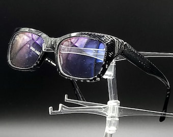 May include: Black rectangular eyeglasses with a textured frame and blue tinted lenses. The glasses are displayed on a clear plastic stand.
