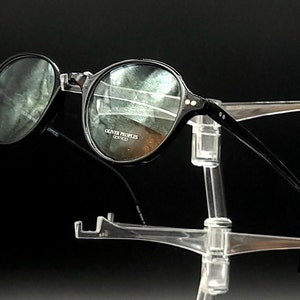 May include: Black round eyeglasses with clear lenses and a black frame. The brand name "Oliver Peoples" is printed on the frame.