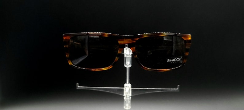 Puede incluir: Gafas de sol de carey con lentes negros. Las gafas tienen el logotipo "SHARCK" en la lente derecha.