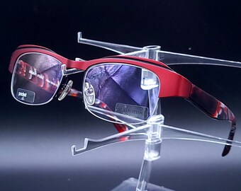 May include: Red-framed eyeglasses with a patterned design on the temples and arms. The lenses reflect the surrounding environment. The glasses are displayed on a clear plastic stand, showcasing their design and style. The glasses have the text "pocket" on the lens.