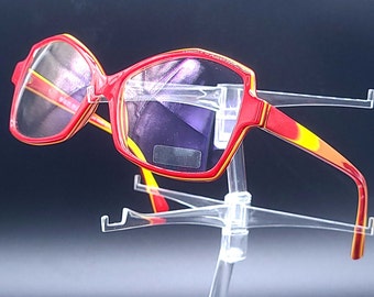 May include: Red and yellow patterned eyeglasses with a clear plastic frame. The glasses are on a clear plastic stand.