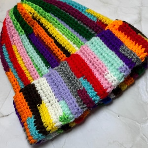 May include: A colorful, handmade crochet beanie hat. The hat features a patchwork design with vertical stripes in various colors, including red, orange, yellow, green, blue, purple, and gray. The brim is folded up, revealing the interior color pattern.
