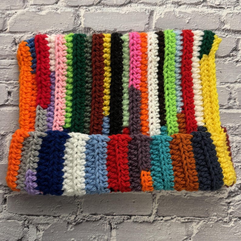 Pu&ograve; includere: Un cappello all'uncinetto fatto a mano e colorato. Il cappello presenta strisce verticali in una variet&agrave; di colori, tra cui rosso, arancione, giallo, verde, blu e viola. Il bordo &egrave; ripiegato e il cappello sembra essere fatto di filato morbido.