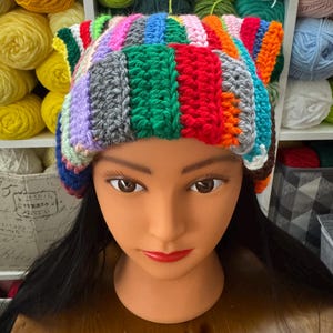 Pu&ograve; includere: Un cappello all'uncinetto colorato e fatto a mano con un design patchwork. Il cappello presenta una variet&agrave; di colori tra cui rosso, verde, blu e arancione. Il cappello &egrave; esposto su una testa di manichino con capelli neri e rossetto rosso.