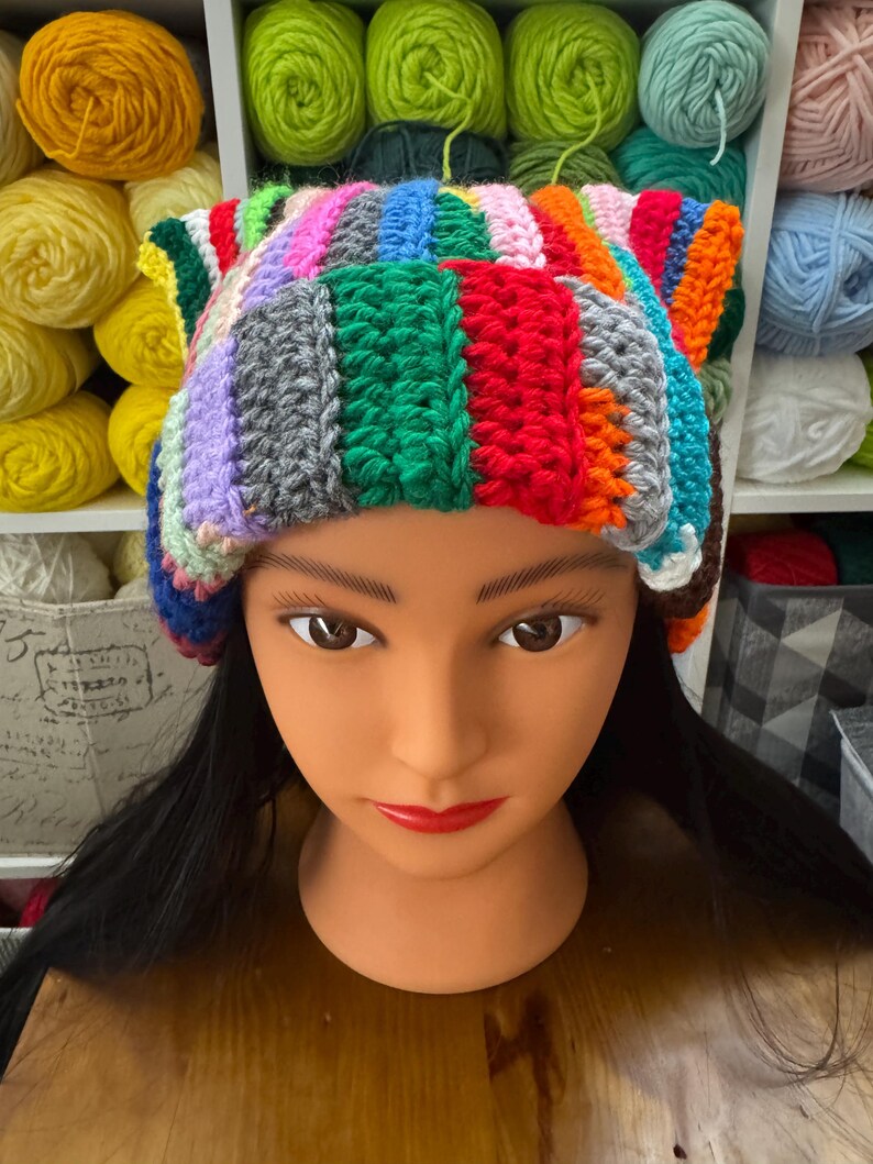 Pu&ograve; includere: Un cappello all'uncinetto colorato e fatto a mano con un design patchwork. Il cappello presenta una variet&agrave; di colori tra cui rosso, verde, blu e arancione. Il cappello &egrave; esposto su una testa di manichino con capelli neri e rossetto rosso.
