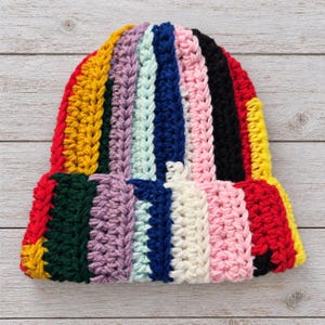 May include: A beanie hat crafted from thick, chunky yarn in a vertical striped pattern. The colors include red, yellow, dark green, purple, light blue, navy blue, pink, black, and cream, featuring a folded brim and textured knit.