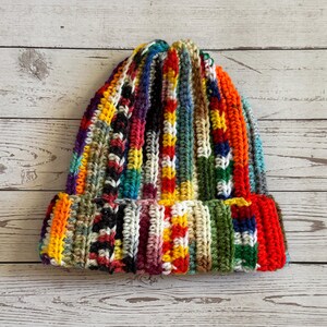 May include: A handmade, colorful crochet beanie hat. The hat features a ribbed design with vertical stripes of various colors, including red, orange, yellow, green, blue, and purple. The brim is folded up.