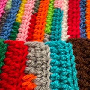Pu&ograve; includere: Primo piano di una colorata coperta all'uncinetto fatta a mano. La coperta presenta strisce verticali in vari colori, tra cui rosa, blu, giallo, arancione, verde, bianco, rosso, grigio, turchese e marrone. La trama &egrave; ben visibile, mostrando le singole maglie.