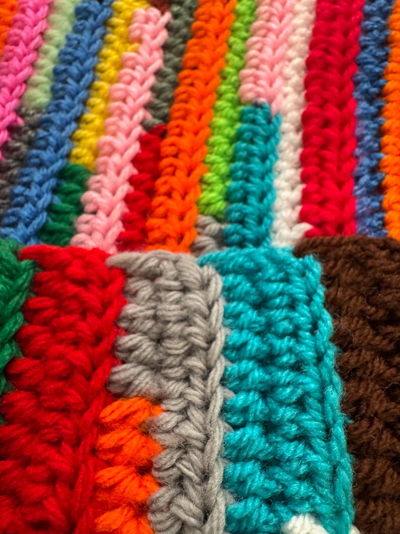 Pu&ograve; includere: Primo piano di una colorata coperta all'uncinetto fatta a mano. La coperta presenta strisce verticali in vari colori, tra cui rosa, blu, giallo, arancione, verde, bianco, rosso, grigio, turchese e marrone. La trama &egrave; ben visibile, mostrando le singole maglie.