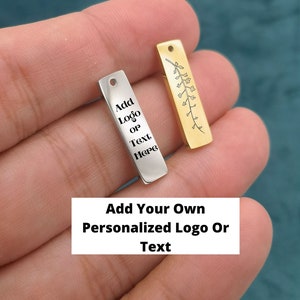 Op de afbeelding: Twee rechthoekige metalen hangers. Eén is zilverkleurig met de tekst "Add Logo or Text Here". De andere is goudkleurig met een bloemenontwerp. De afbeelding bevat ook de tekst "Add Your Own Personalized Logo Or Text".