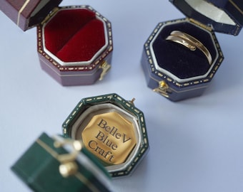 May include: Three ring boxes, one red, one blue and one green. The green box is open and shows a gold ring with the inscription "Belle V Blue Craft".