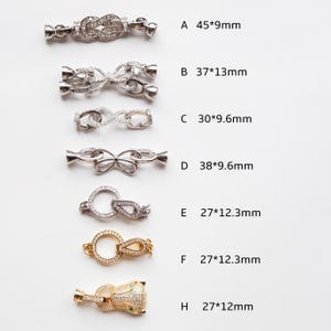 Könnte beinhalten: Sieben silberne und goldfarbene Metallverschlüsse mit klaren Kristallverzierungen. Die Verschlüsse sind in verschiedenen Formen und Größen, darunter ein Kreis, ein Unendlichkeitszeichen und eine Schleife. Die Maße jedes Verschlusses sind neben dem Buchstabenlabel angegeben.