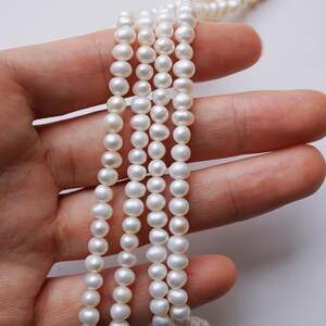 May include: A strand of white pearl beads, strung on a thin thread, is held in a hand. The pearls are round and smooth, and the strand is long enough to wrap around a wrist or neck.