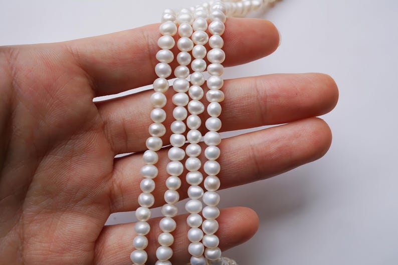 May include: A strand of white pearl beads, strung on a thin thread, is held in a hand. The pearls are round and smooth, and the strand is long enough to wrap around a wrist or neck.