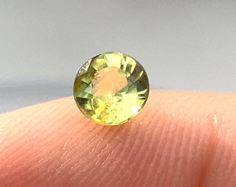 May include: A round, faceted gemstone with a pale yellow-green hue. The gem is held between two fingers, showcasing its clarity and cut. The gemstone appears to be a peridot, a type of olivine, often used in jewelry.