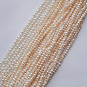 Op de afbeelding: Een streng witte en crèmekleurige zoetwaterparels. De parels zijn klein en rond en zijn aan elkaar geregen op een dun draad. De parels zijn gerangschikt in een aflopend patroon, met de grootste parels bovenaan en de kleinste parels onderaan.