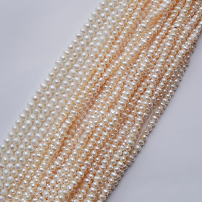 May include: A strand of white and cream-colored freshwater pearls. The pearls are small and round, and they are strung together on a thin thread. The pearls are arranged in a graduated pattern, with the largest pearls at the top and the smallest pearls at the bottom.