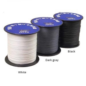 Puede incluir: Tres carretes de hilo de pescar en blanco, gris oscuro y negro. Cada carrete tiene una etiqueta azul con texto japonés y las palabras "300M JAPÓN".