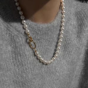 May include: A pearl necklace with a gold clasp. The necklace features a strand of white pearls and a gold-colored clasp with an oval design. The necklace is worn against a gray sweater.